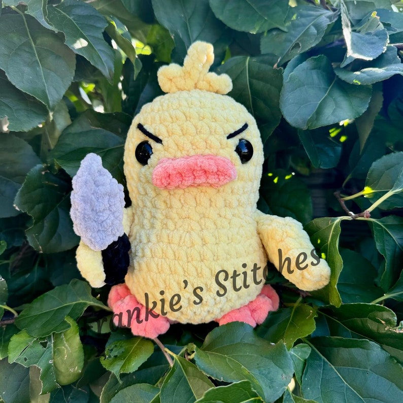 Puede incluir: Un pato amarillo de ganchillo hecho a mano con ojos negros, pico rosa y pies rosas, sosteniendo un cuchillo gris y negro. El texto "Frankie's Stitches" es visible en la parte inferior derecha. El pato est&aacute; sobre un fondo de hojas verdes.