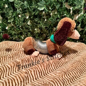 May include: A handmade dachshund dog toy with a brown and burgundy crochet body, a silver slinky midsection, and a green collar. The dog is lying on a brown, textured surface. The text "Frankie's Stitches" is visible.