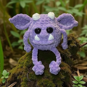 May include: A handmade, plush purple monster with large black eyes, white horns, and white fangs. The creature has large ears and is sitting on a mossy surface. The plush toy is a whimsical, soft sculpture.