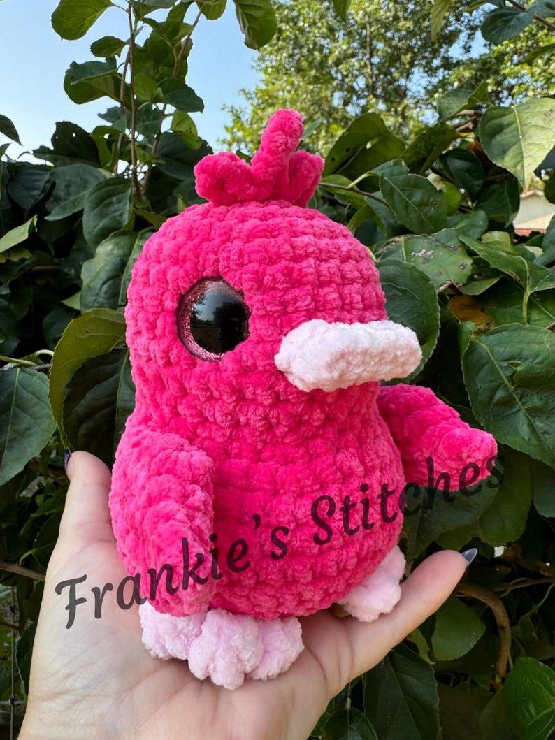 Puede incluir: Un p&aacute;jaro rosa de peluche hecho a mano, con grandes ojos negros, un pico rosa claro y patas a juego. El p&aacute;jaro tiene un aspecto texturizado y difuso y una peque&ntilde;a cresta. El texto "Frankie's Stitches" es visible en el p&aacute;jaro.