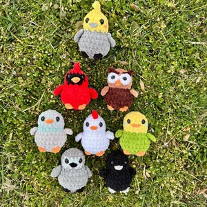 May include: A collection of nine crocheted animal plushies, including a cardinal, owl, chick, penguin, and more. The plushies are arranged on a bed of green grass.