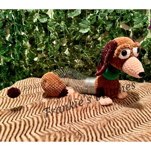 May include: A handmade crochet dachshund dog toy in brown, burgundy, and tan. The toy has large, expressive eyes, a green collar, and a spring-like body. The toy is on a brown and tan patterned surface.