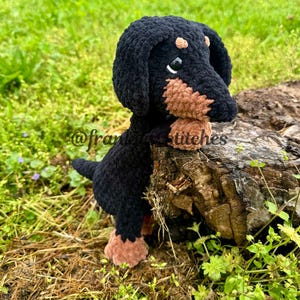 May include: A black and brown crocheted dachshund toy with large eyes, sitting on a tree stump. The toy is leaning against the stump and has a sad expression.