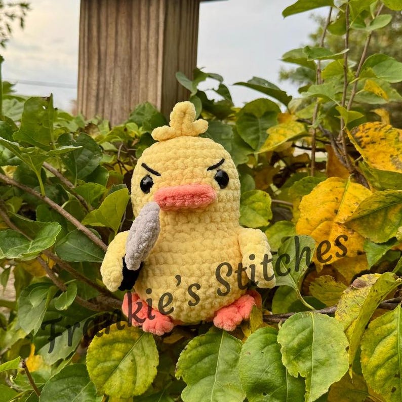 Puede incluir: Un pato amarillo de ganchillo hecho a mano con una expresi&oacute;n enfadada, sosteniendo un cuchillo gris. El pato tiene un pico y patas de color melocot&oacute;n y ojos negros. El texto "Frankie's Stitches" es visible en el lado derecho de la imagen.