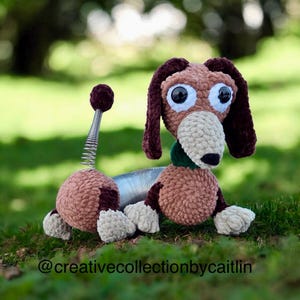 May include: A handmade crochet dachshund dog toy in shades of brown, tan, and burgundy, with large, expressive eyes. The dog features a spring-like midsection and a green collar, resting on green grass. The text "@creativecollectionbycaitlin" is at the bottom.