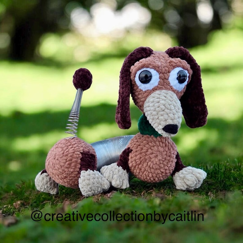 May include: A handmade crochet dachshund dog toy in shades of brown, tan, and burgundy, with large, expressive eyes. The dog features a spring-like midsection and a green collar, resting on green grass. The text "@creativecollectionbycaitlin" is at the bottom.