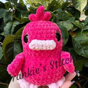 Puede incluir: Una criatura de crochet rosa brillante hecha a mano, con forma de p&aacute;jaro, con grandes ojos negros, un pico blanco y peque&ntilde;os pies rosas. El texto "Frankie's Stitches" est&aacute; impreso en el lateral. El peluche est&aacute; sobre un fondo de hojas verdes.