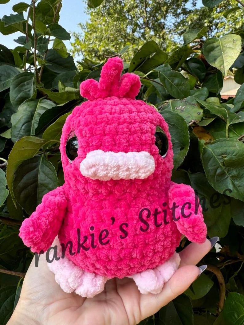Puede incluir: Una criatura de crochet rosa brillante hecha a mano, con forma de p&aacute;jaro, con grandes ojos negros, un pico blanco y peque&ntilde;os pies rosas. El texto "Frankie's Stitches" est&aacute; impreso en el lateral. El peluche est&aacute; sobre un fondo de hojas verdes.