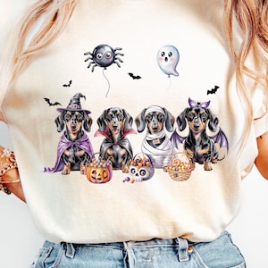 May include: Cream-colored t-shirt featuring a Halloween-themed graphic. The design includes five dachshunds dressed in costumes, such as a witch, vampire, and mummy. A spider balloon and ghost balloon float above the dogs, with bats scattered around.