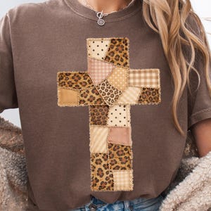 Puede incluir: Una camiseta marrón con un diseño de cruz de retazos. La cruz está compuesta por varios patrones, incluyendo estampado de leopardo, lunares y cuadros, en tonos beige, rosa y marrón. La camiseta tiene cuello redondo y mangas cortas.