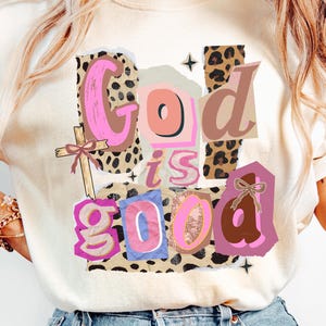 May include: A cream-coloured t-shirt featuring the phrase "God is Good" in a collage design. The design incorporates pink, brown, and leopard print elements, plus a cross graphic. A casual and inspirational apparel item.