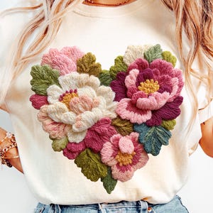 Könnte beinhalten: Cremefarbenes T-Shirt mit einem herzförmigen Blumendesign. Die gestickten Blumen weisen Farbtöne von Rosa, Weiß und Burgunder auf, mit grünen Blättern und gelben Zentren. Das Shirt hat kurze Ärmel und einen Rundhalsausschnitt.