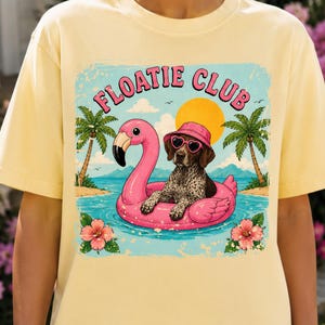 May include: Yellow t-shirt featuring a graphic of a dog wearing sunglasses and a hat, floating on a pink flamingo floatie. The graphic includes the words "Floatie Club", palm trees, and a sun over a beach scene.