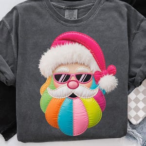 May include: A dark gray sweatshirt featuring a colorful Santa Claus face with sunglasses. The Santa face is made up of various colors, including pink, orange, blue, and green. The Santa hat is pink with white trim, and a pink bow is on the side.