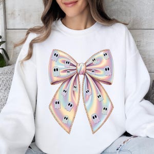May include: White sweatshirt featuring a large, colorful bow design. The bow has a pastel rainbow gradient with gold trim and is adorned with small, cartoon ghost illustrations. A festive and whimsical garment.
