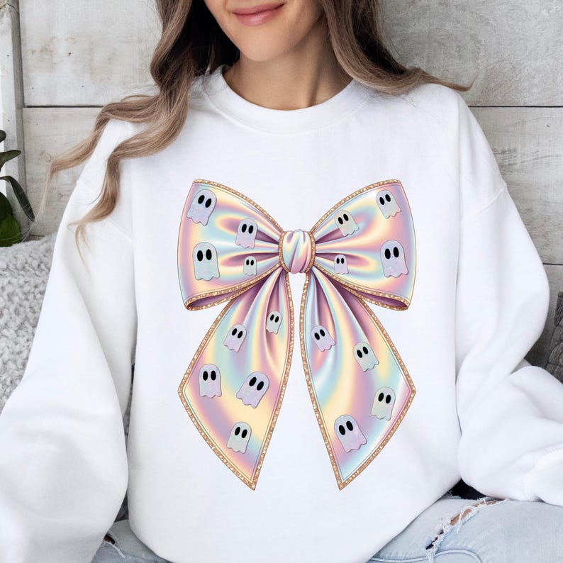 May include: White sweatshirt featuring a large, colorful bow design. The bow has a pastel rainbow gradient with gold trim and is adorned with small, cartoon ghost illustrations. A festive and whimsical garment.