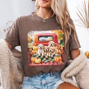 Pode incluir: Camiseta marrom com uma imagem de uma caminhonete vermelha cheia de abóboras e cinco filhotes de golden retriever. A caçamba da caminhonete tem um cobertor xadrez. O fundo mostra uma cena de outono com árvores e folhagens.