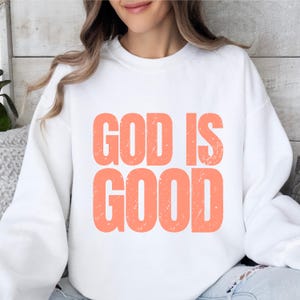 May include: White sweatshirt with the text "GOD IS GOOD" in a coral color. The text has a distressed, vintage look. The sweatshirt is a casual, comfortable style.