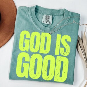 May include: A seafoam green t-shirt with the words "GOD IS GOOD" in bright yellow, distressed lettering. The shirt is folded and has a Comfort Colors tag. A silver necklace and a brown hat are visible.