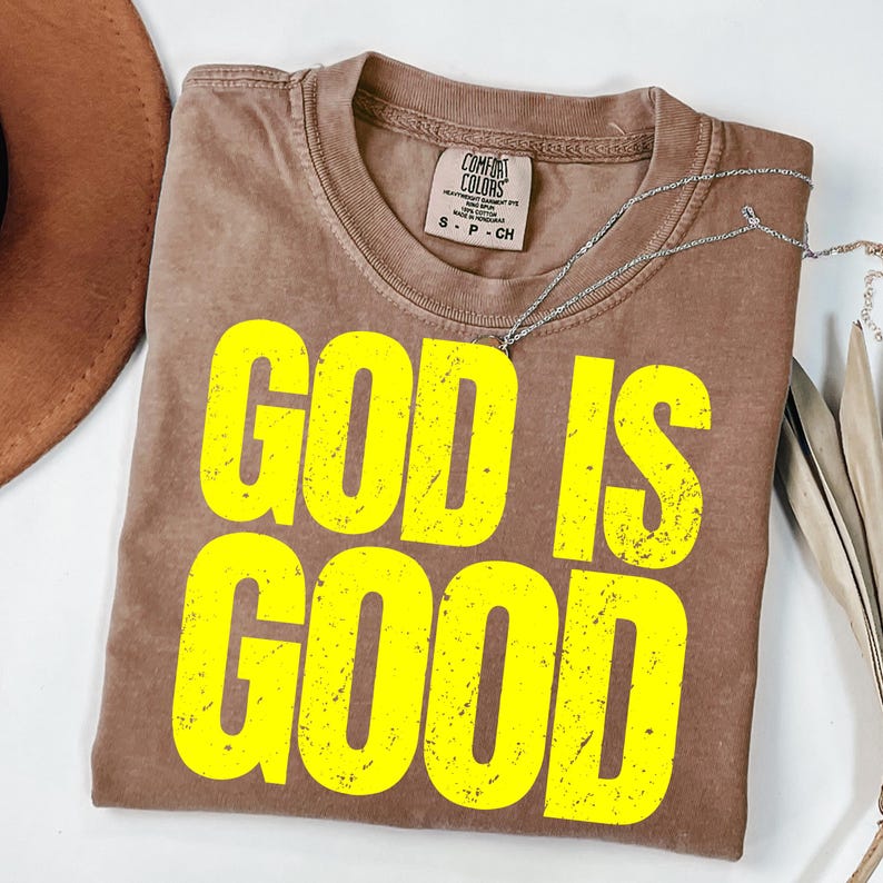 May include: A brown Comfort Colors t-shirt with the words "GOD IS GOOD" in bright yellow block letters. A silver necklace is draped across the shirt. The shirt is folded and lies on a white surface.