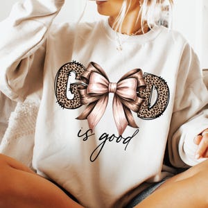 May include: Cream-colored sweatshirt with the words "GOD" in a leopard print design, a large pink bow, and the phrase "is good" in cursive. The sweatshirt is a casual style.