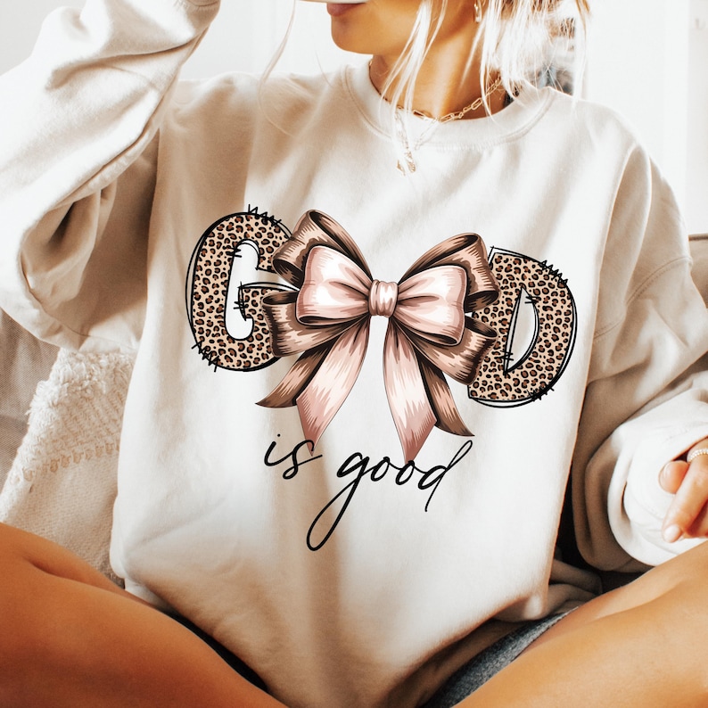 May include: Cream-colored sweatshirt with the words "GOD" in a leopard print design, a large pink bow, and the phrase "is good" in cursive. The sweatshirt is a casual style.