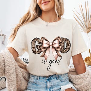 May include: Cream-colored t-shirt with the words "GOD is good" in a script font. The letters "G" and "D" are filled with a leopard print design, and a large pink bow is centered between the letters.