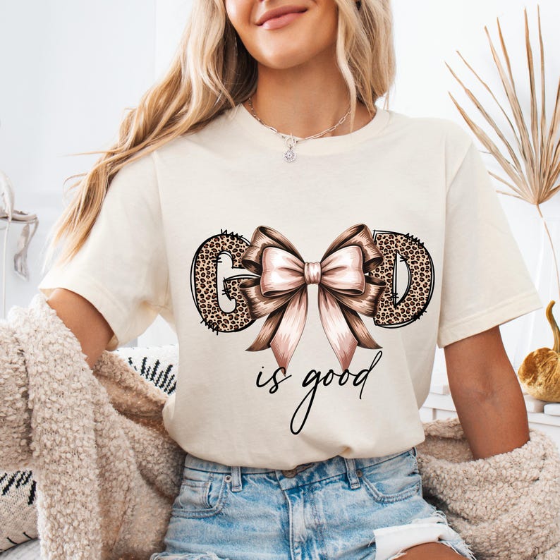 May include: Cream-colored t-shirt with the words "GOD is good" in a script font. The letters "G" and "D" are filled with a leopard print design, and a large pink bow is centered between the letters.