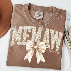 May include: A brown t-shirt with the word "MEMAW" in floral print, a cream-colored bow, and the text "Est. 2026". The shirt has a Comfort Colors tag and a silver necklace. A brown hat is visible in the upper left corner.