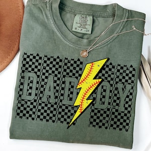 Op de afbeelding: Saliegroen t-shirt met het woord "DADDY" in een zwart-wit geblokt patroon. Een gele bliksemschicht, ontworpen als honkbal, staat in het midden van het shirt. Een zilveren ketting met een kleine hanger ligt op het shirt.