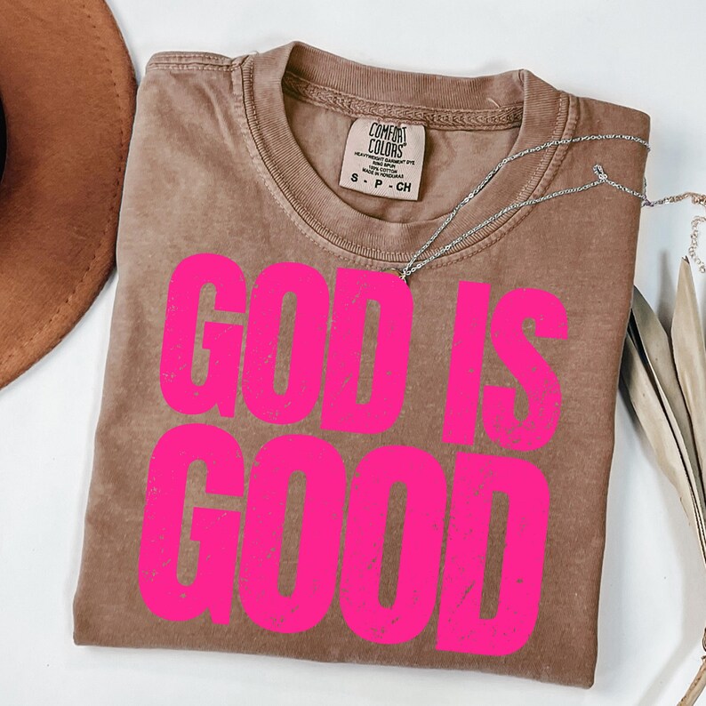 May include: A brown Comfort Colors t-shirt with the words "GOD IS GOOD" in bright pink block letters. A silver necklace is draped across the shirt. A brown hat and decorative leaves are in the background.