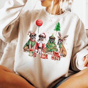 Pu&ograve; includere: Una felpa color crema con un design a tema natalizio. Raffigura quattro cani Boxer vestiti con abiti festivi: un elfo, Babbo Natale, un albero di Natale e una renna. Un palloncino rosso mostra le parole "Merry Christmas".