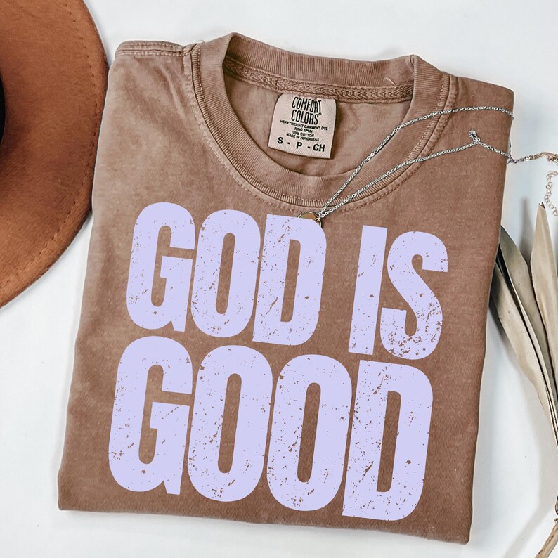 May include: A brown, short-sleeved t-shirt with the words "GOD IS GOOD" in large, light purple letters. The shirt has a crew neck and a small tag at the neckline. A silver necklace is draped across the shirt.
