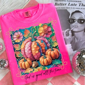 May include: Bright pink Comfort Colors sweatshirt featuring a colorful pumpkin graphic with flowers and the text "God is good all the time." The design includes orange, pink, and teal colors, with gold accents. A newspaper clipping and disco ball are in the background.