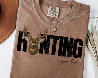 May include: A brown t-shirt with the words "HUNTING Season" in black block letters and script. A detailed illustration of a deer is incorporated into the word "HUNTING". A silver necklace with a small pendant is draped across the shirt.