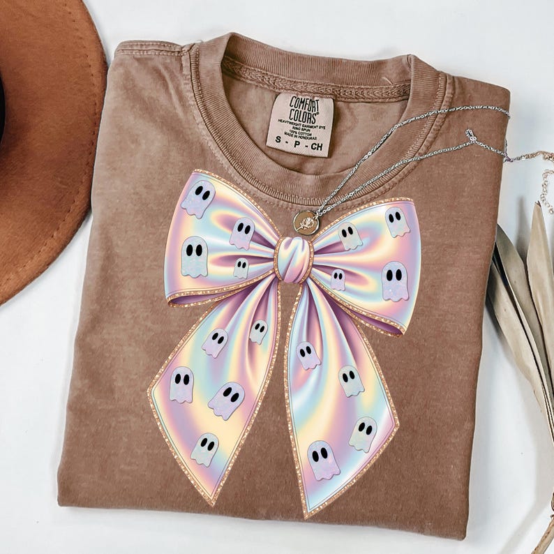 May include: A brown Comfort Colors t-shirt featuring a large, iridescent bow design adorned with cartoon ghosts. The bow has a gold outline and a gradient of pastel colors. A silver necklace with a pendant is draped over the shirt.