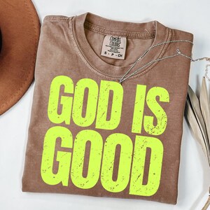 May include: A brown Comfort Colors t-shirt with the words "GOD IS GOOD" in bright yellow. A silver necklace is draped across the shirt. The shirt is folded and placed on a white surface.