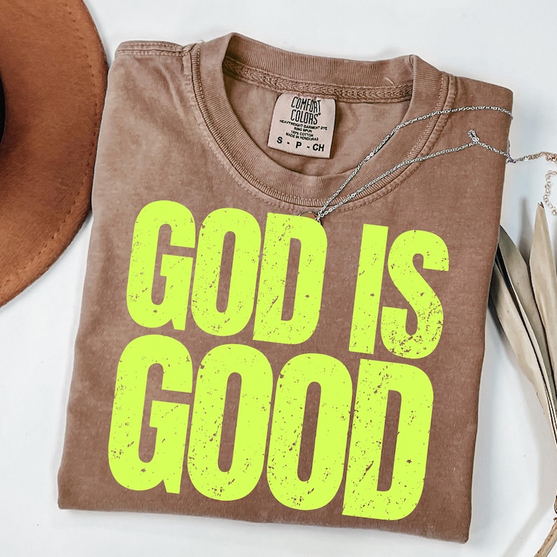 May include: A brown Comfort Colors t-shirt with the words "GOD IS GOOD" in bright yellow. A silver necklace is draped across the shirt. The shirt is folded and placed on a white surface.