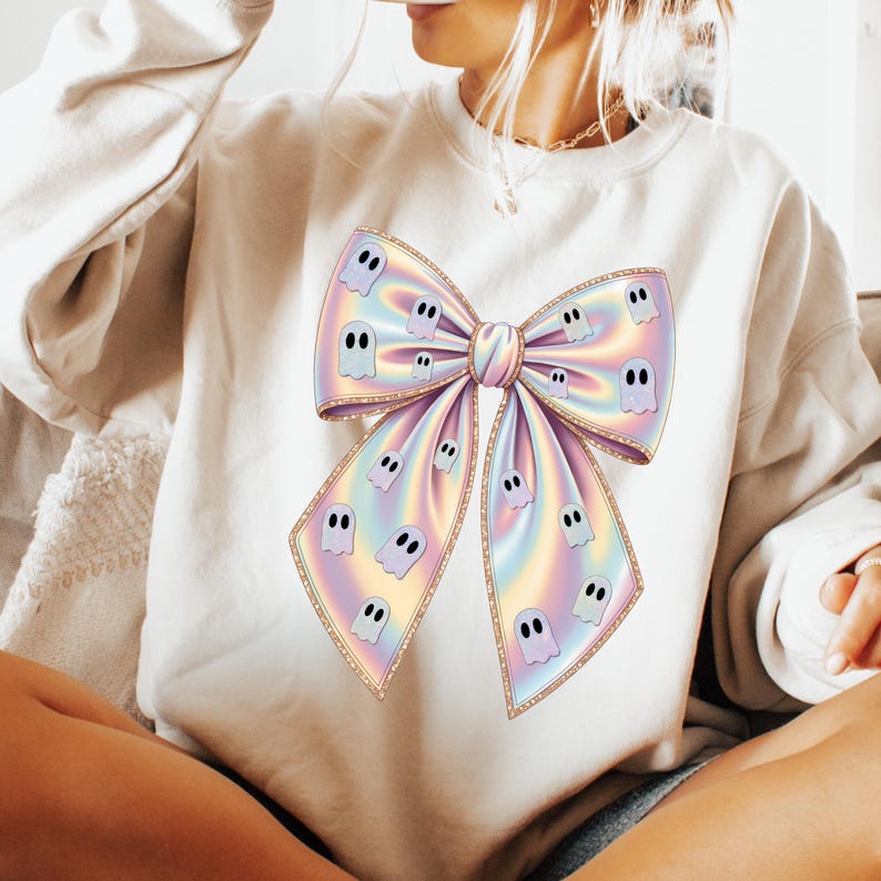 May include: Cream-colored sweatshirt featuring a large, iridescent bow design. The bow is adorned with multiple cartoon ghost figures. The bow has a gold trim. The sweatshirt is a casual, comfortable garment.