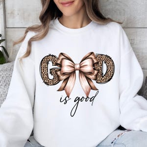 May include: White sweatshirt with the words "GOD" in leopard print and a large, light brown bow. The phrase "is good" is written in a cursive font below the bow. The sweatshirt is a casual, comfortable style.