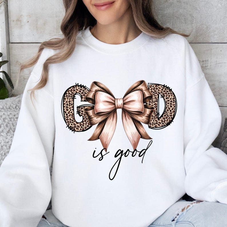 May include: White sweatshirt with the words "GOD" in leopard print and a large, light brown bow. The phrase "is good" is written in a cursive font below the bow. The sweatshirt is a casual, comfortable style.