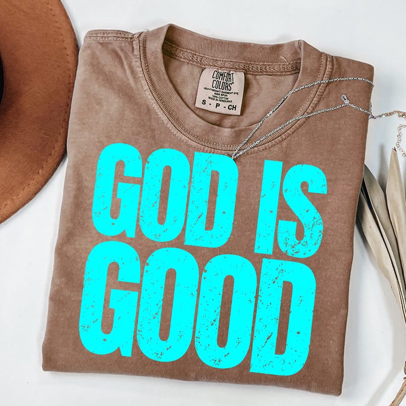 May include: A brown t-shirt with the words "GOD IS GOOD" in bright turquoise letters. The shirt has a crew neck and short sleeves. A silver necklace is draped across the shirt. The shirt is made of a soft material.