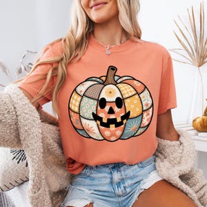 May include: Peach-colored t-shirt featuring a patchwork pumpkin design. The pumpkin has a smiling face and is composed of various fabric patterns in orange, yellow, brown, and blue. A fall-themed apparel item.