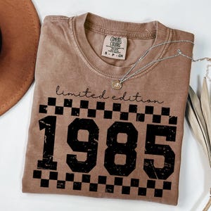 May include: A brown t-shirt with a black checkered pattern and the text "limited edition 1985" in a distressed font.