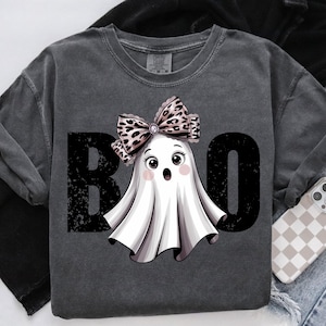 Può includere: Felpa grigio scuro con grafica a tema Halloween. Il design presenta un fantasma dei cartoni animati con un fiocco leopardato e la scritta "BOO" in nero sbiadito. È visibile un telefono con una custodia a scacchi.