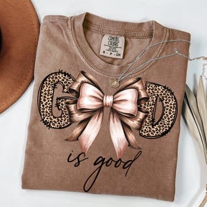 May include: A brown t-shirt with the words "GOD is good" in a script font. The letters "G" and "D" are filled with a leopard print design. A large pink bow is in the centre of the design. A silver necklace with a small pendant is on the shirt.