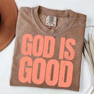 May include: A brown Comfort Colors t-shirt with the words "GOD IS GOOD" in coral lettering. A silver necklace is draped across the shirt. The shirt is folded on a white surface.