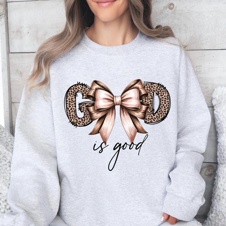 May include: Light gray sweatshirt with the words "GOD is good" in a script font. The letters "G" and "D" are leopard print. A large bow with brown and pink ribbons is centered above the words.