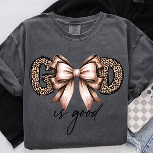 May include: Dark gray sweatshirt with the words "GOD is good" in a script font. The letters "GOD" are filled with a leopard print design, and a large bow with pink and brown ribbons is centered above the words.