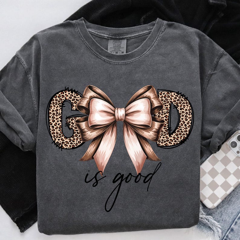 May include: Dark gray sweatshirt with the words "GOD is good" in a script font. The letters "GOD" are filled with a leopard print design, and a large bow with pink and brown ribbons is centered above the words.